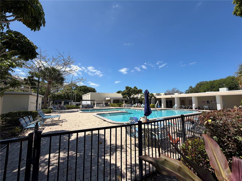 A fenced outdoor swimming pool area with lounge chairs and surrounding buildings under a blue sky — 5967 TERRACE PARK DRIVE N UNIT 104, ST PETERSBURG, FL