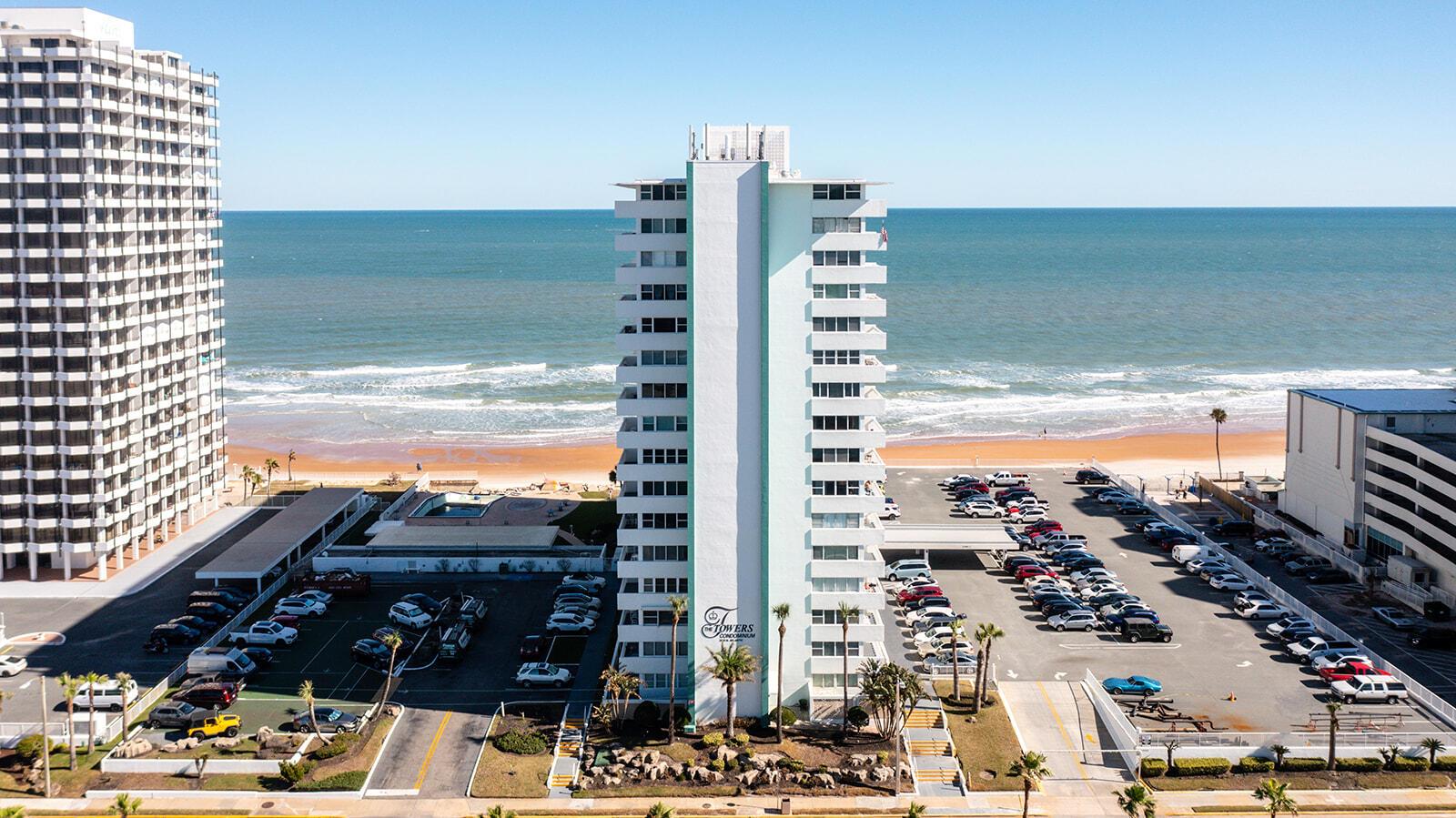 An aerial view shows a tall, light-colored condominium building situated between a parking lot and the beach — 2800 N Atlantic Avenue Unit 712, FL 32118, FL