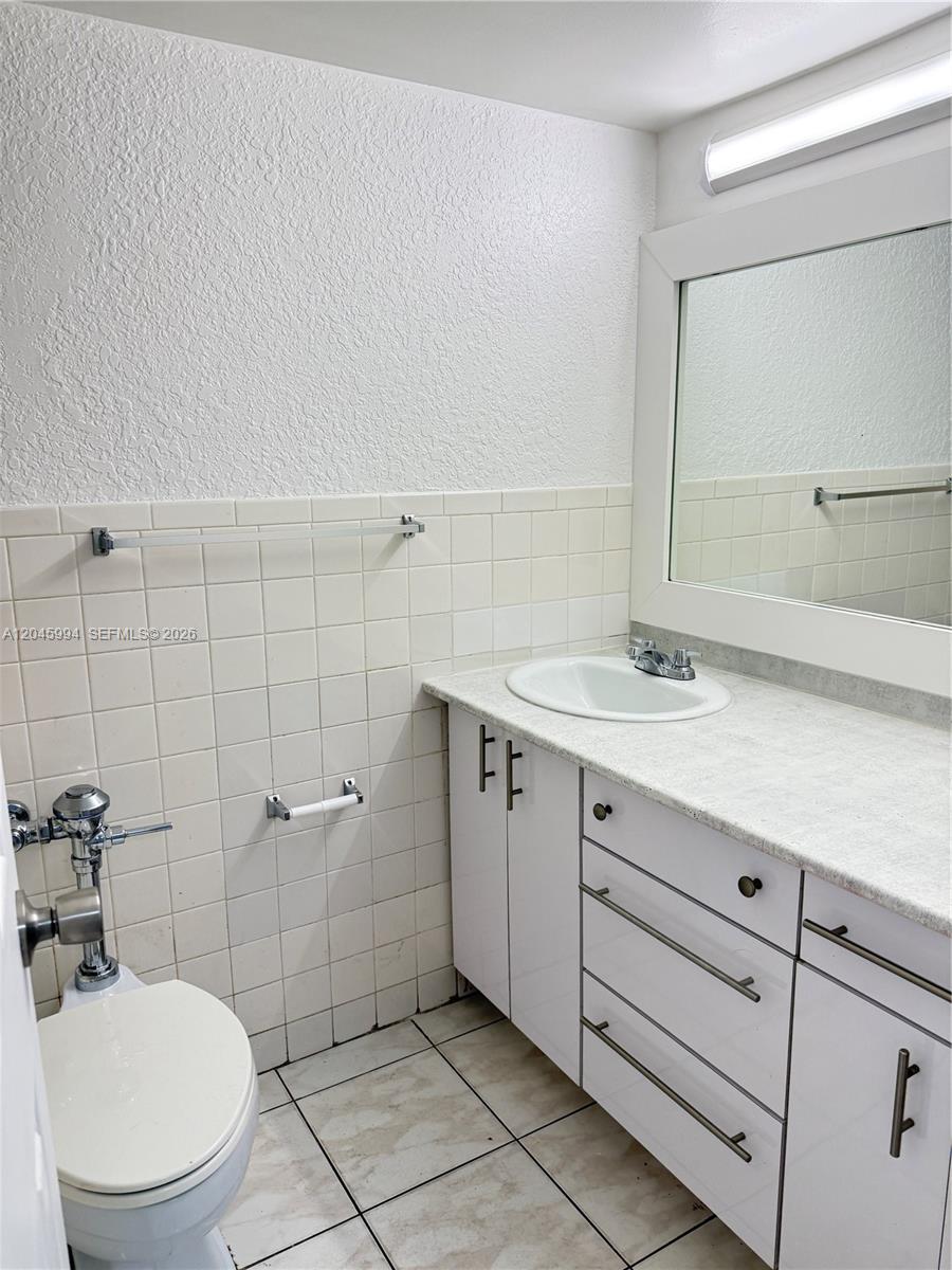 A bathroom features a white vanity, a wall-mounted mirror, a toilet, and tiled walls — HALLANDALE BEACH, FL