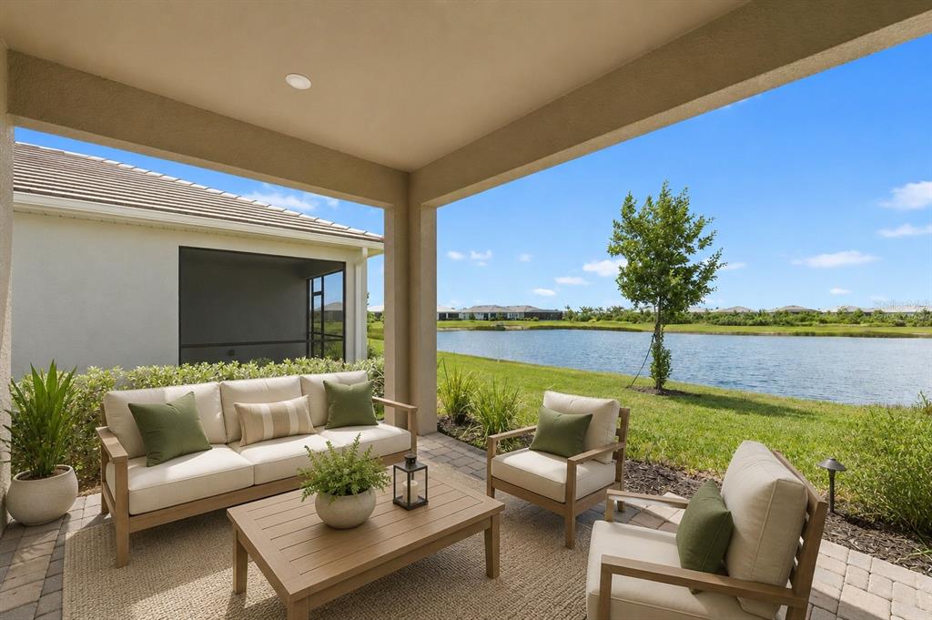 A covered patio features outdoor furniture overlooking a grassy lawn and a lake — 11566 BOUNDLESS TERRACE, VENICE, FL