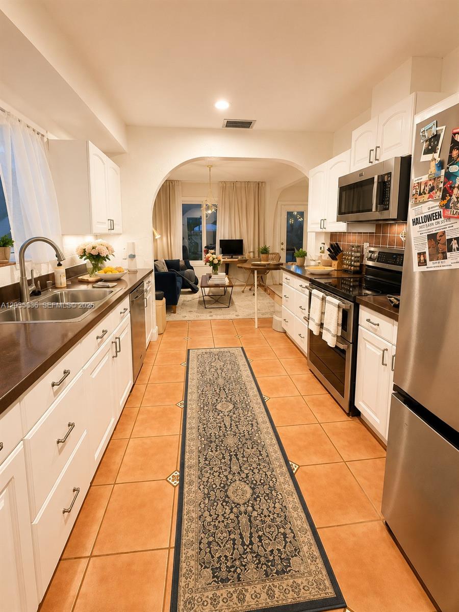 A galley kitchen with white cabinets and tile floors leads into a living area — 1183 NE 88th St, Miami FL 33138, MIAMI, FL