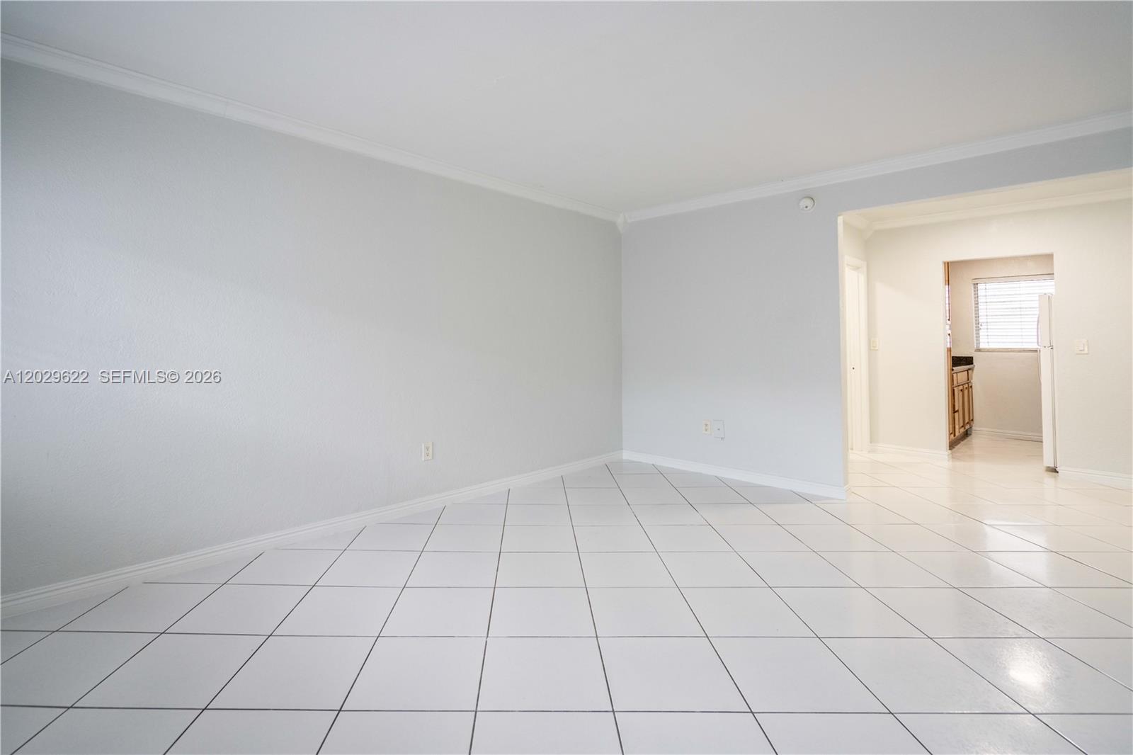 A bright, empty room features white tiled floors, light gray walls, and a doorway leading to a kitchen — 7640 Carlyle Ave # 9B, Miami Beach FL 33141, MIAMI BEACH, FL