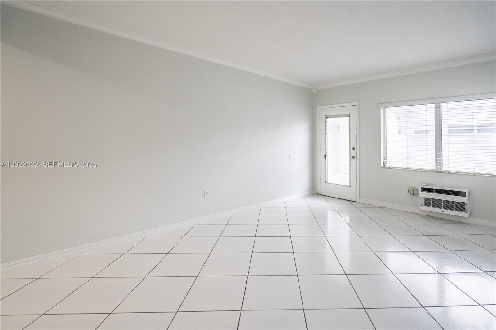 An empty room features white tiled floors, light gray walls, a window, and a door — 7640 Carlyle Ave # 9B, Miami Beach FL 33141, MIAMI BEACH, FL