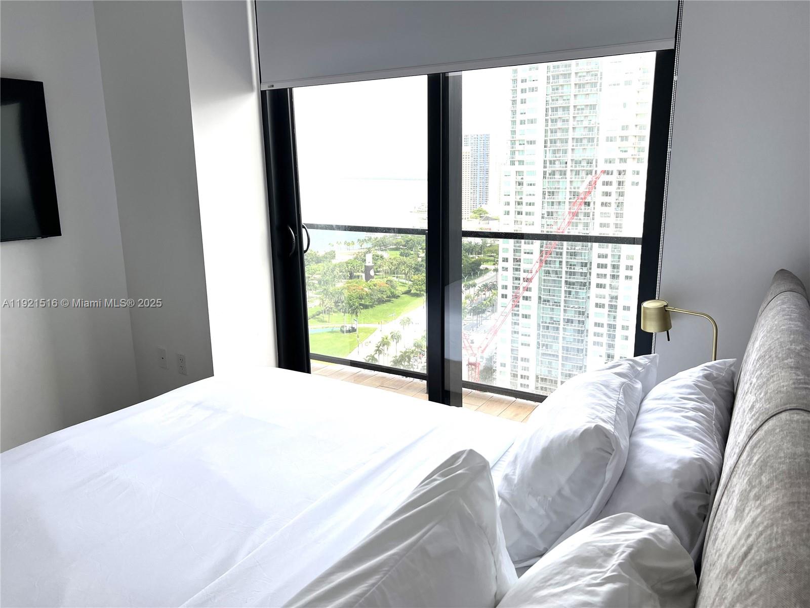 A bedroom features a bed with white linens and a large window overlooking city buildings — 398 NE 5th St # 2215, Miami FL 33132, MIAMI, FL