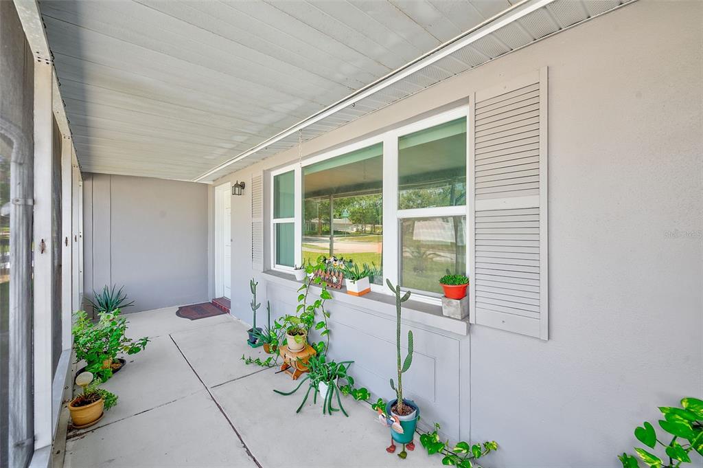 A covered concrete porch features a large window with shutters and several potted plants — 5126 ROSEWOOD DRIVE, NEW PORT RICHEY, FL