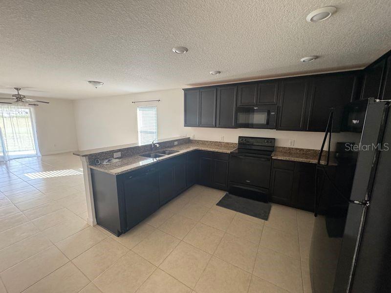 A kitchen with dark cabinets, granite countertops, and black appliances opens to a living area — 6909 CRESTED ORCHID DRIVE, BROOKSVILLE, FL