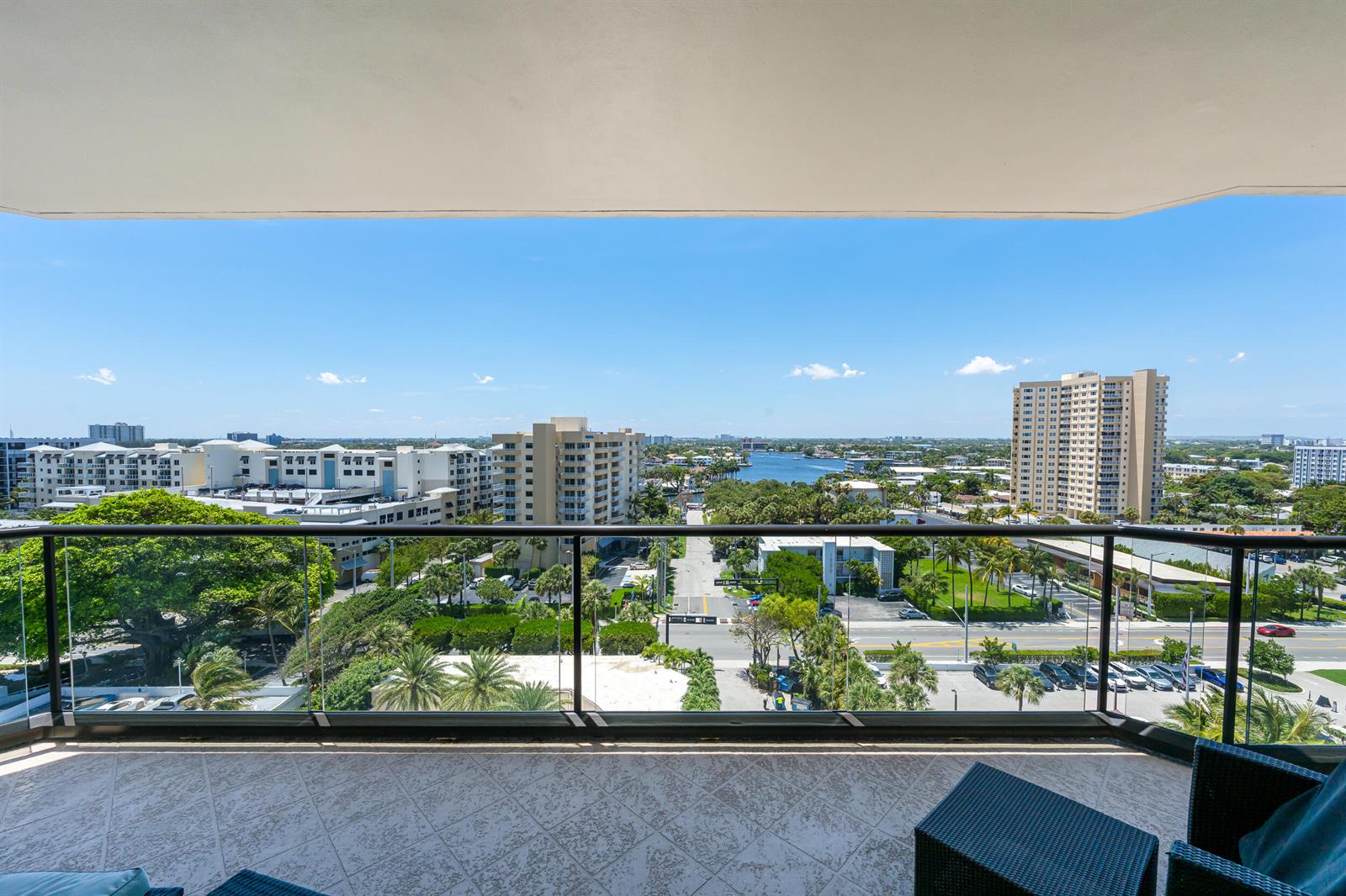 A balcony with a glass railing overlooks a city street, palm trees, and distant water — 1300 S Ocean Boulevard # 906, Pompano Beach FL 33062, POMPANO BEACH, FL