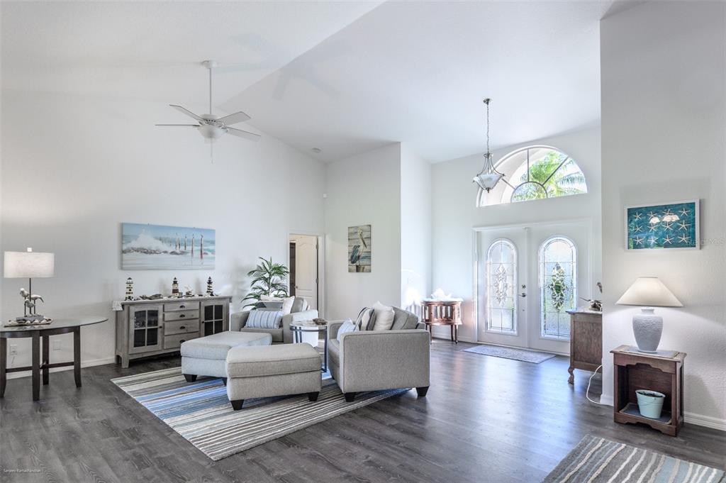A bright living room features vaulted ceilings, gray wood-look flooring, and a double-door entryway — 27406 TIERRA DEL FUEGO CIRCLE, PUNTA GORDA, FL
