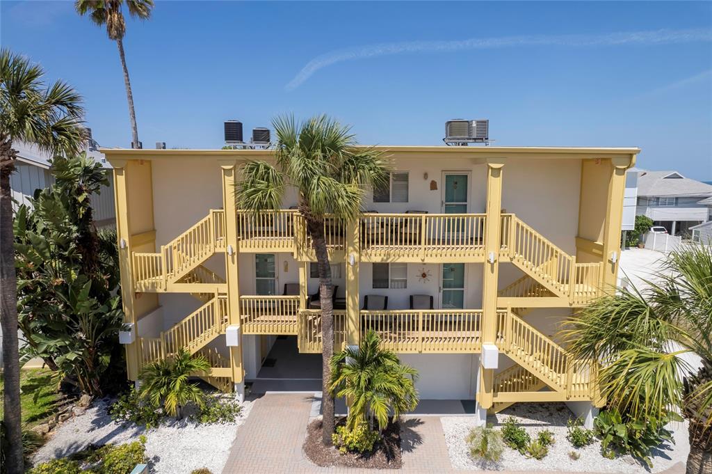 A three-story yellow building features exterior staircases, balconies, and palm trees in a sunny setting — 708 GULF BOULEVARD UNIT D, INDIAN ROCKS BEACH, FL