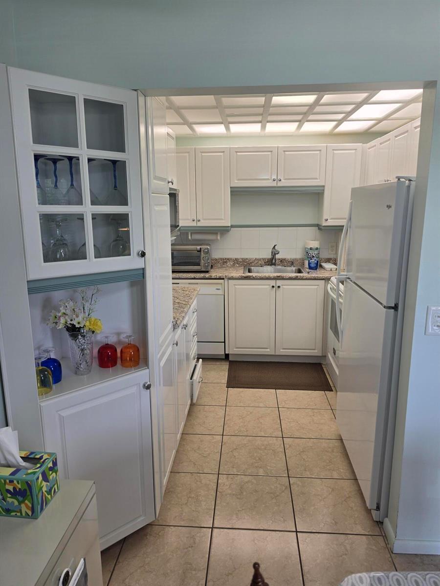 A galley kitchen features white cabinets, light countertops, tile flooring, and a glass-front display cabinet — 15 Markham # A, Deerfield Beach FL 33442, DEERFIELD BEACH, FL