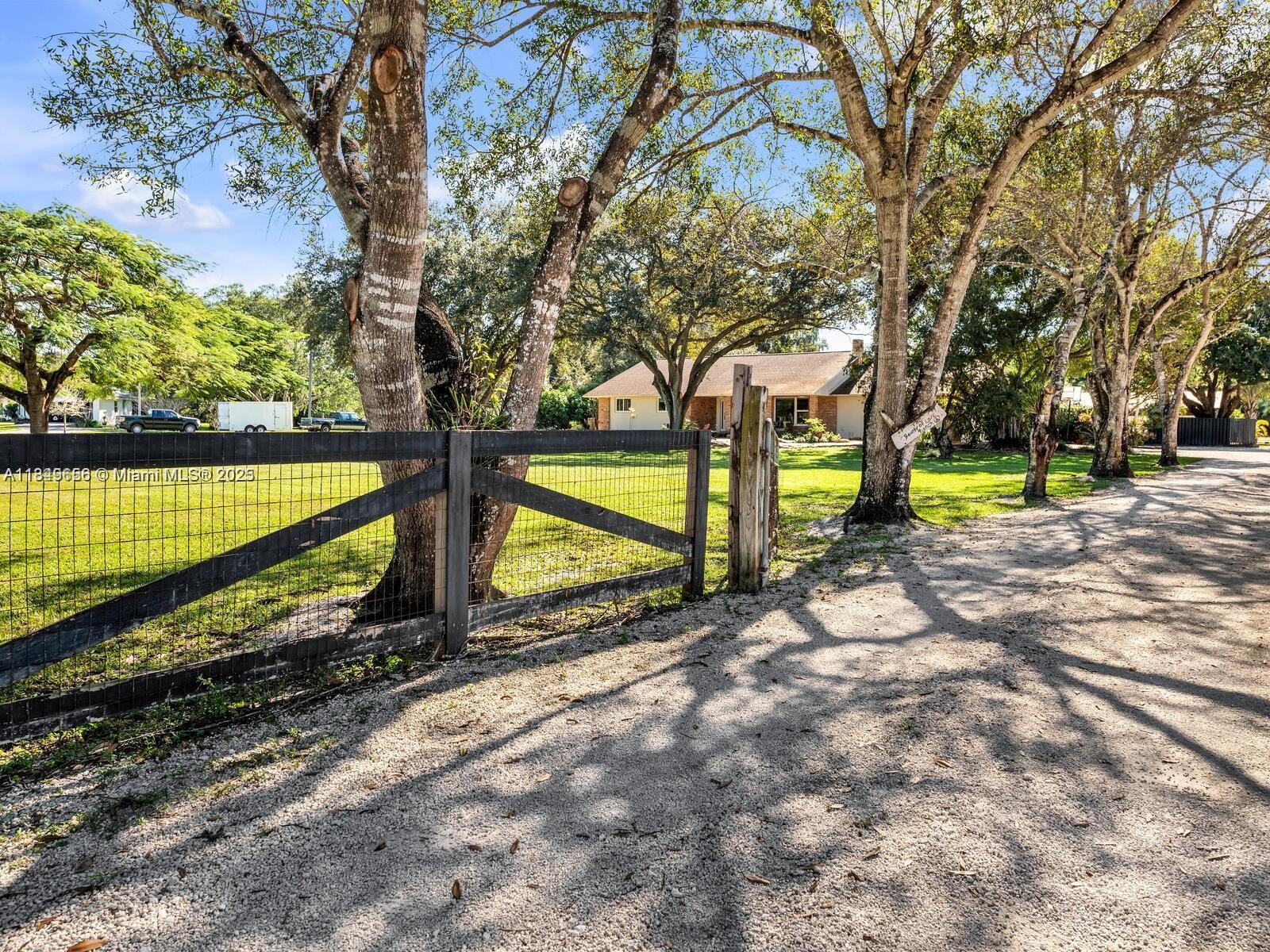 A gravel driveway leads toward a house behind a wooden fence and large trees — 13350 Luray Rd, Southwest Ranches FL 33330, SOUTHWEST RANCHES, FL