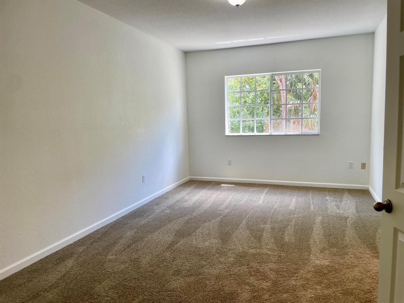 An empty bedroom features beige carpet, white walls, and a window overlooking green trees — 5064 SE Mariner Garden Circle, Stuart FL 34997, STUART, FL