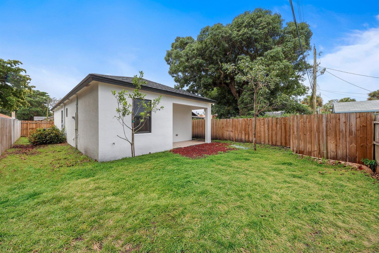 A white stucco house with a small covered patio sits in a fenced grassy backyard — 633 55th Street, West Palm Beach FL 33407, WEST PALM BEACH, FL