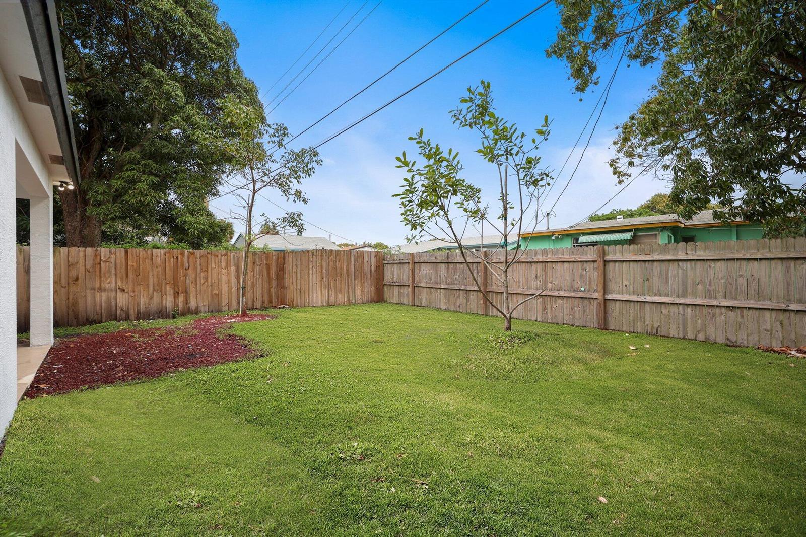 A fenced backyard features a grassy lawn, two young trees, and a patch of mulch — 633 55th Street, West Palm Beach FL 33407, WEST PALM BEACH, FL
