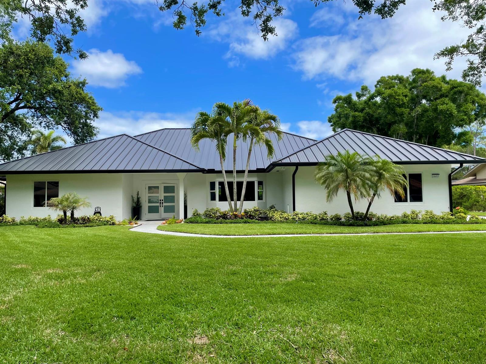 A white single-story house with a dark metal roof and palm trees in the front yard — 18457 SE Heritage Oaks Lane, Jupiter FL 33469, JUPITER, FL