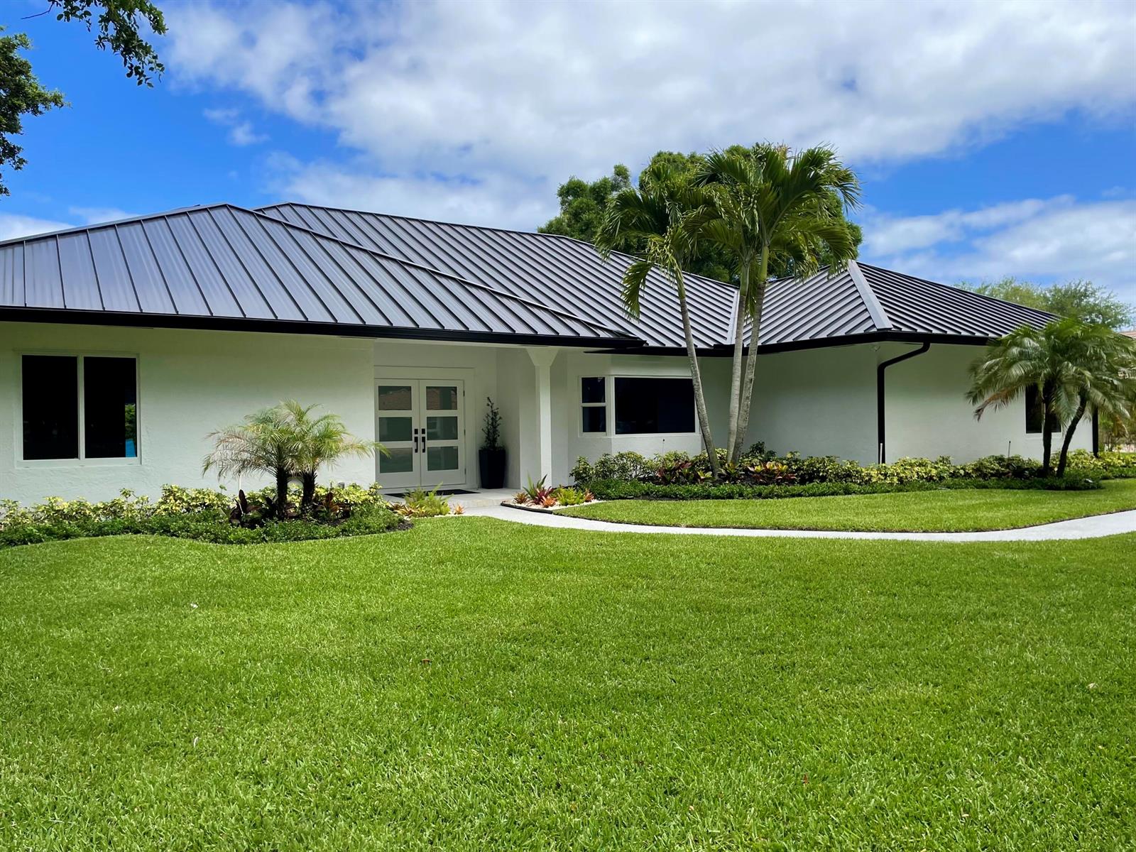 A white single-story house with a dark metal roof and palm trees on a lawn — 18457 SE Heritage Oaks Lane, Jupiter FL 33469, JUPITER, FL