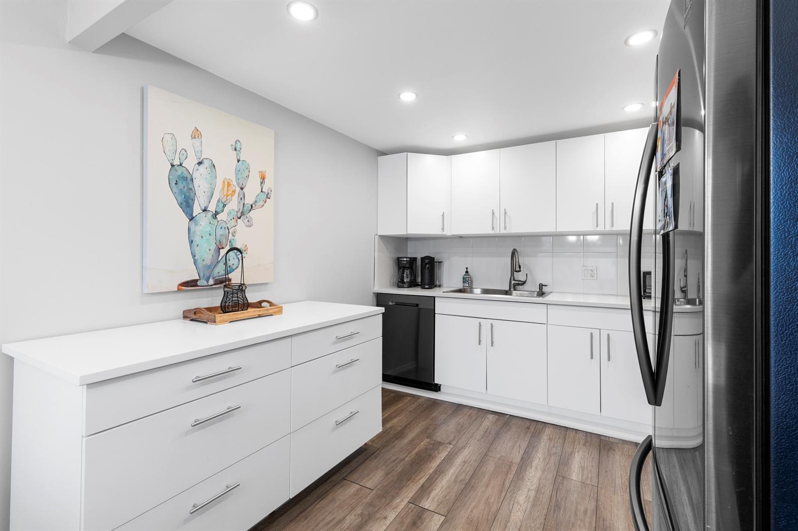 A modern kitchen features white cabinets, a black dishwasher, and a cactus wall art piece — 1032 Ventnor H Crescent # H, Deerfield Beach FL 33442, DEERFIELD BEACH, FL