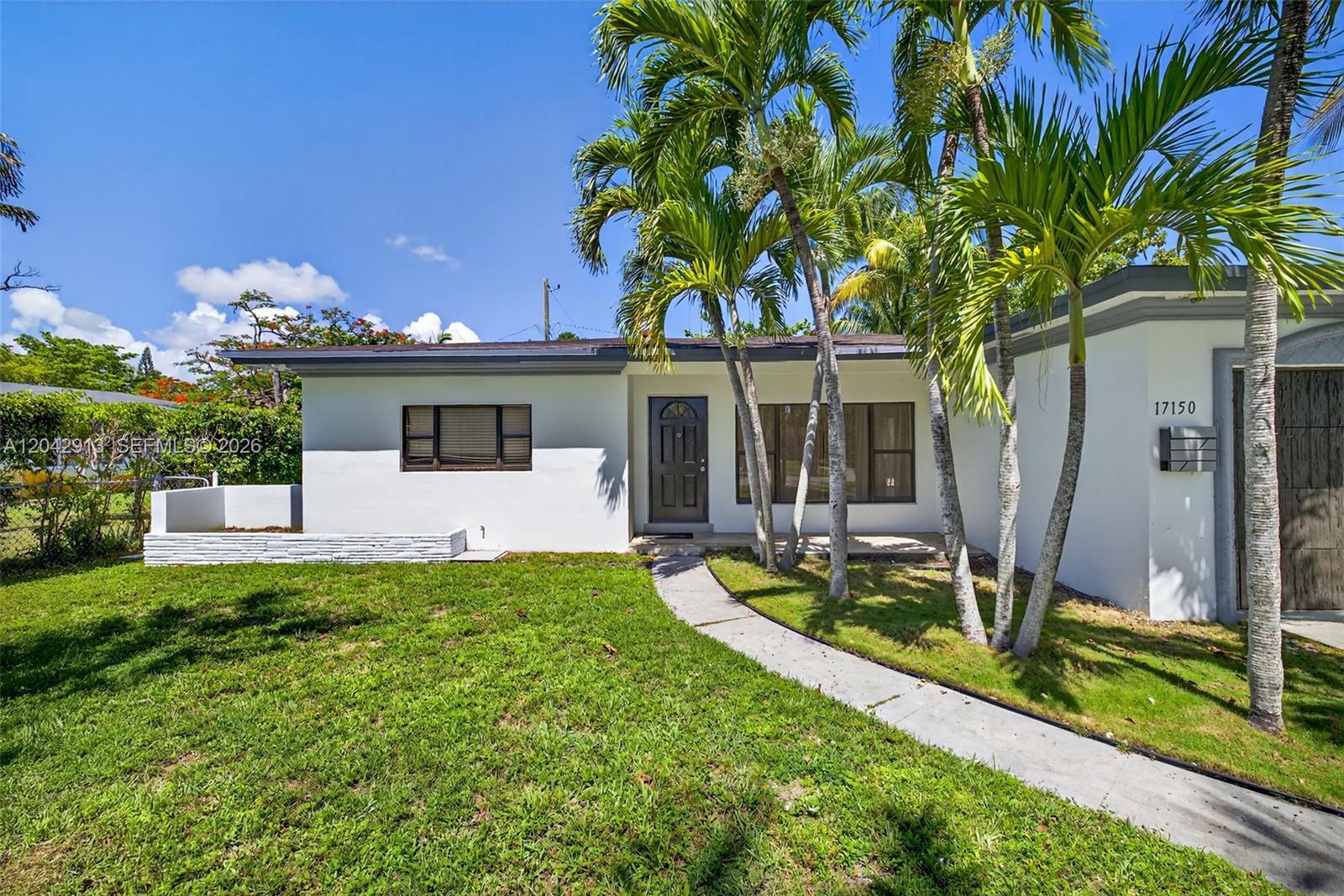 A white single-story house features a front walkway, palm trees, and a small stone wall — NORTH MIAMI BEACH, FL