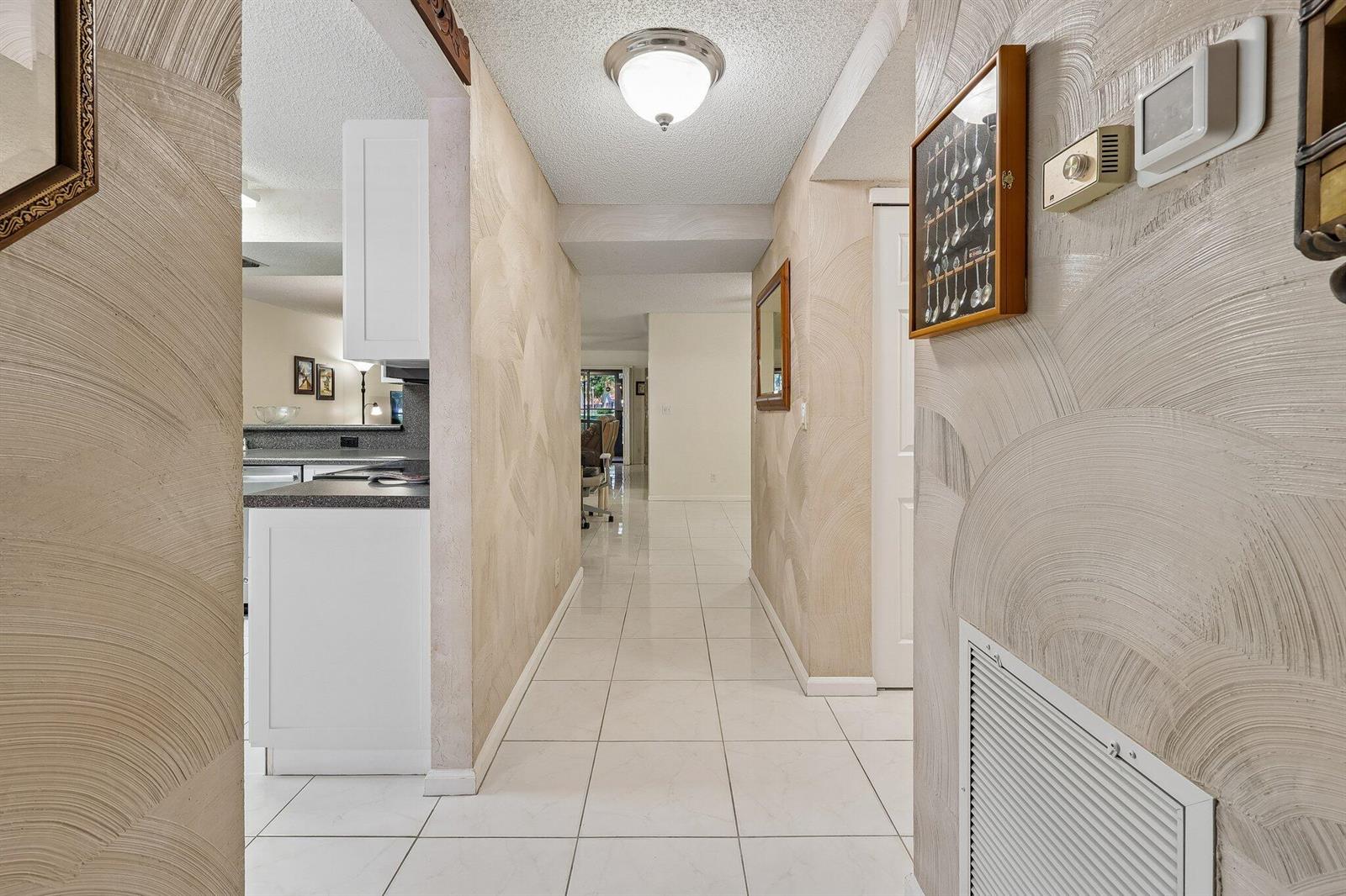 A hallway with textured beige walls, white tile flooring, and a ceiling-mounted light fixture — 9681 E Sills Dr E # 102, Boynton Beach FL 33437, BOYNTON BEACH, FL