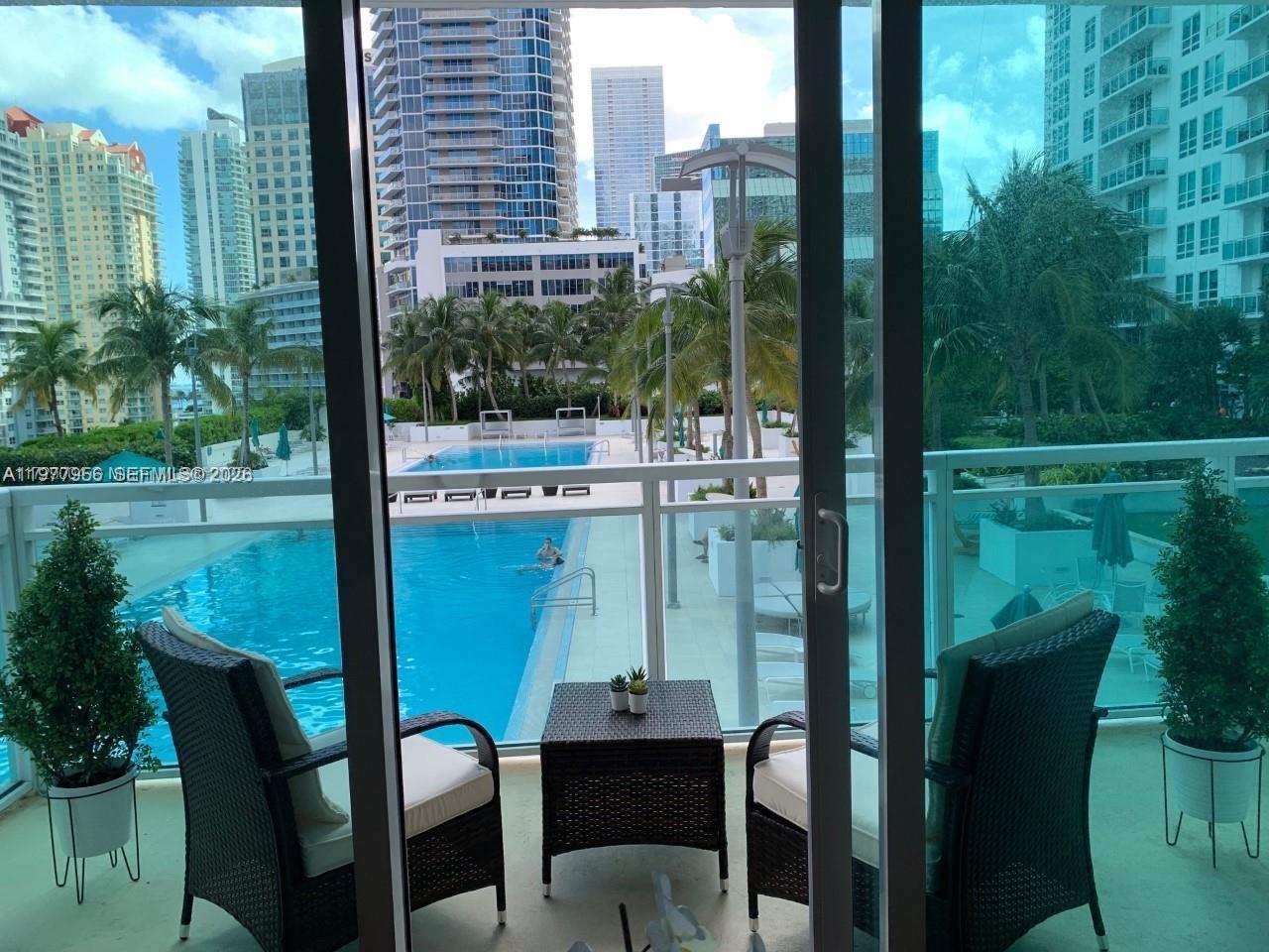 A balcony with two chairs and a table overlooks a swimming pool and city buildings — 950 Brickell Bay Dr # 1107, Miami FL 33131, MIAMI, FL