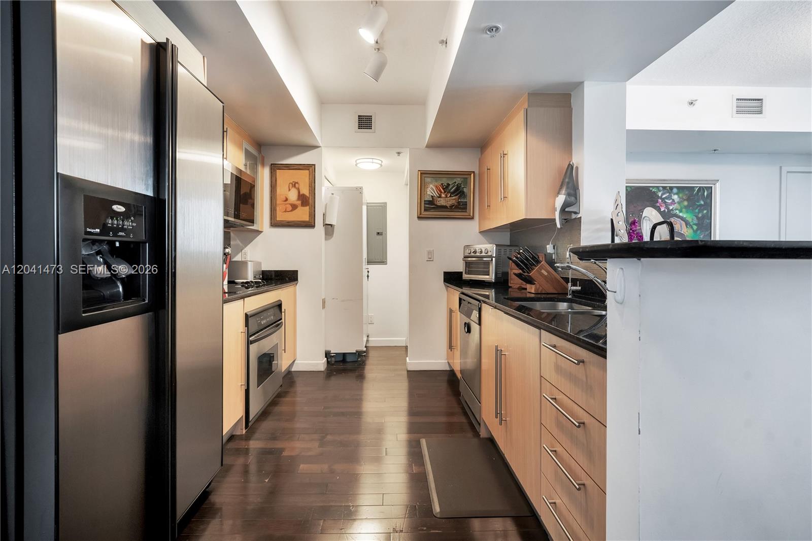 A galley kitchen features light wood cabinets, black countertops, stainless steel appliances, and dark flooring — 325 S Biscayne Blvd # 3921, Miami FL 33131, MIAMI, FL