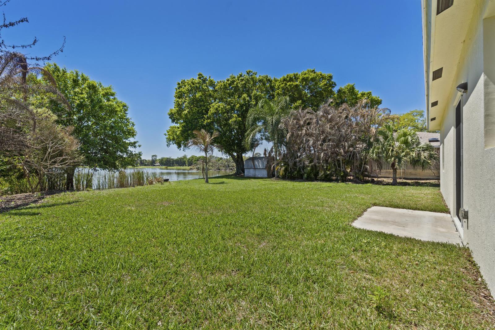 A grassy backyard features a small concrete patio, mature trees, and a view of a lake — 7705 Sebastian Road, Fort Pierce FL 34951, FORT PIERCE, FL