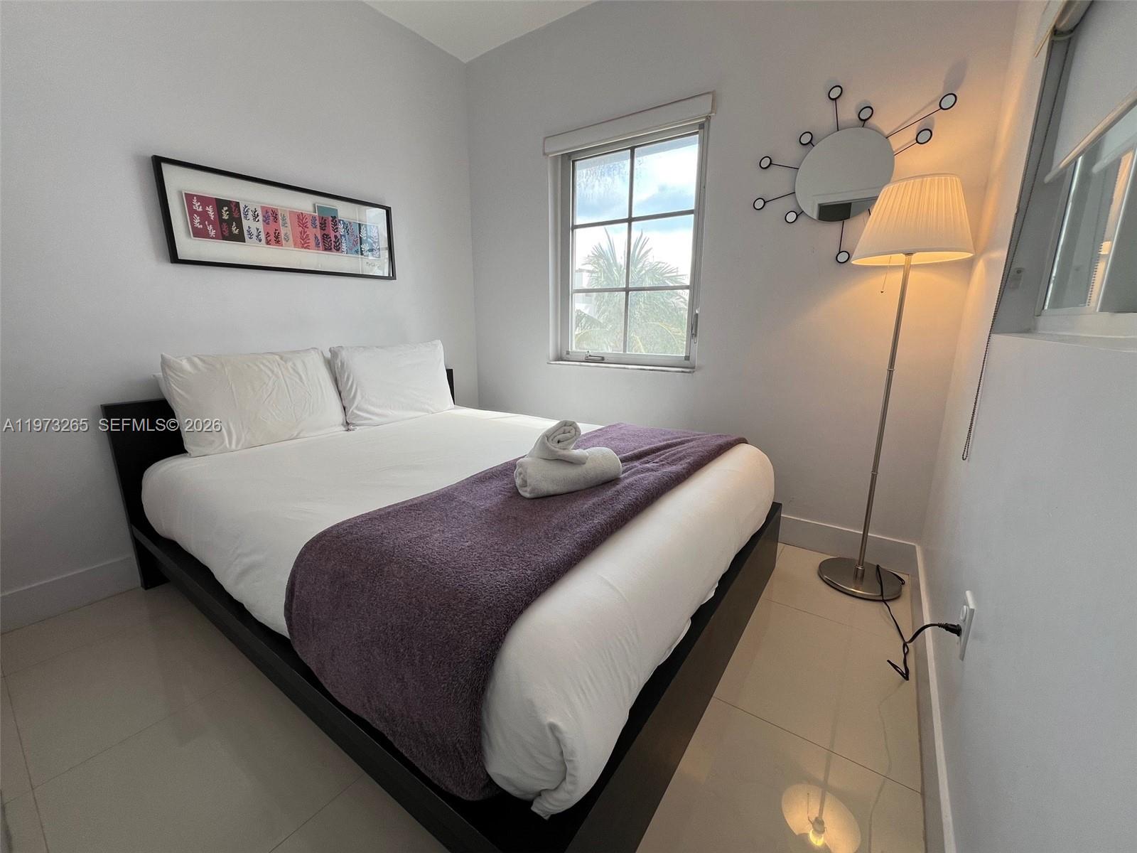 A bedroom features a bed with a purple throw, a floor lamp, and wall art — 1100 Collins Ave # 303, Miami Beach FL 33139, MIAMI BEACH, FL