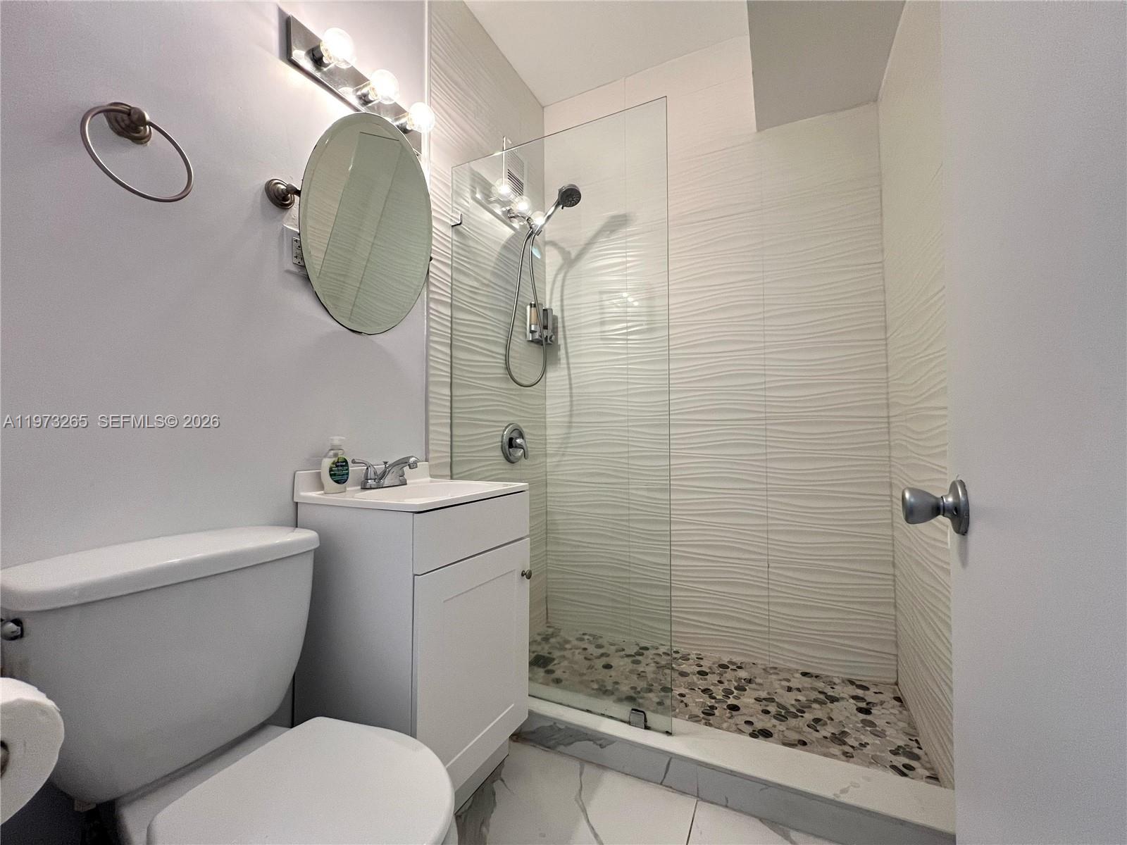 A small bathroom features a white vanity, a toilet, and a glass-enclosed pebble-floor shower — 1100 Collins Ave # 303, Miami Beach FL 33139, MIAMI BEACH, FL