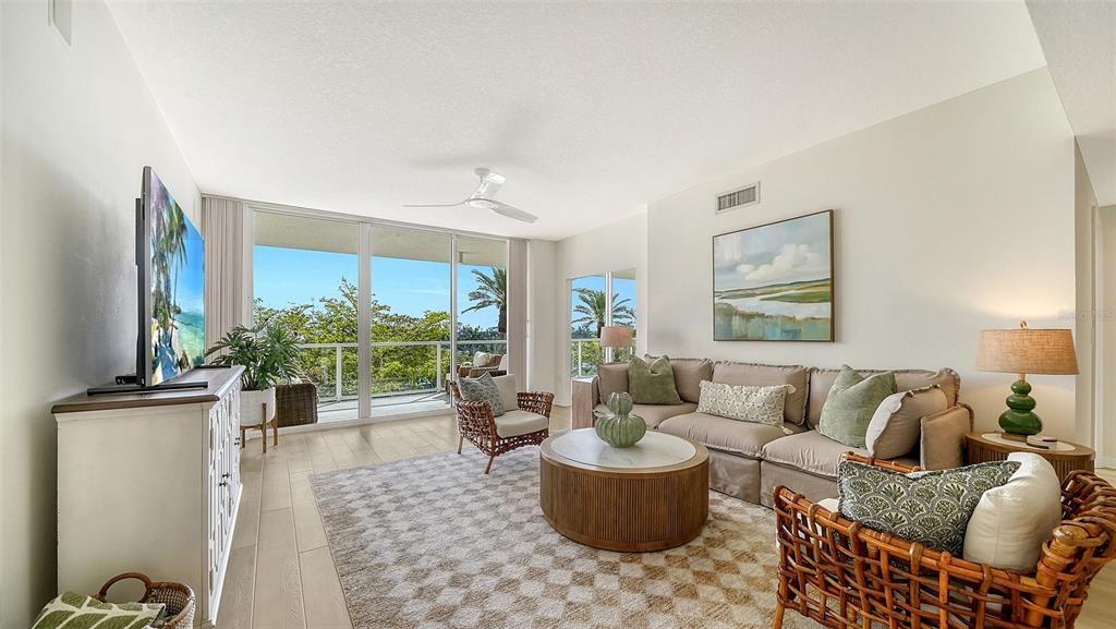 A bright living room features a beige sofa, rattan chairs, and a balcony with greenery — 1800 BENJAMIN FRANKLIN DRIVE UNIT B308, SARASOTA, FL