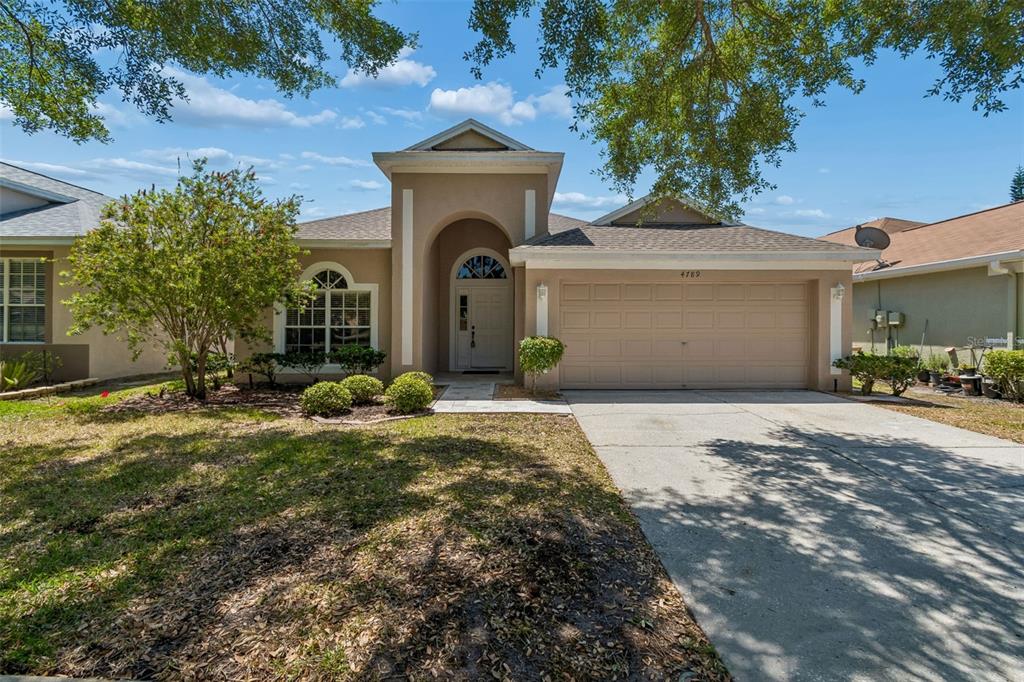 A single-story suburban house features a beige stucco exterior, a two-car garage, and arched entryway — 4789 WHISPERING WIND AVENUE, TAMPA, FL