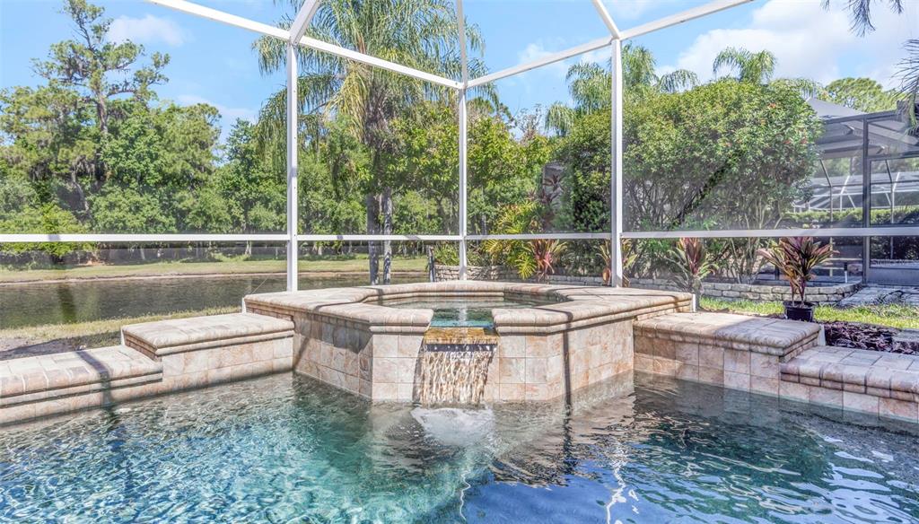 A tiled spa with a spillover waterfall sits inside a screened pool enclosure by water — 4152 GRANDCHAMP CIRCLE, PALM HARBOR, FL