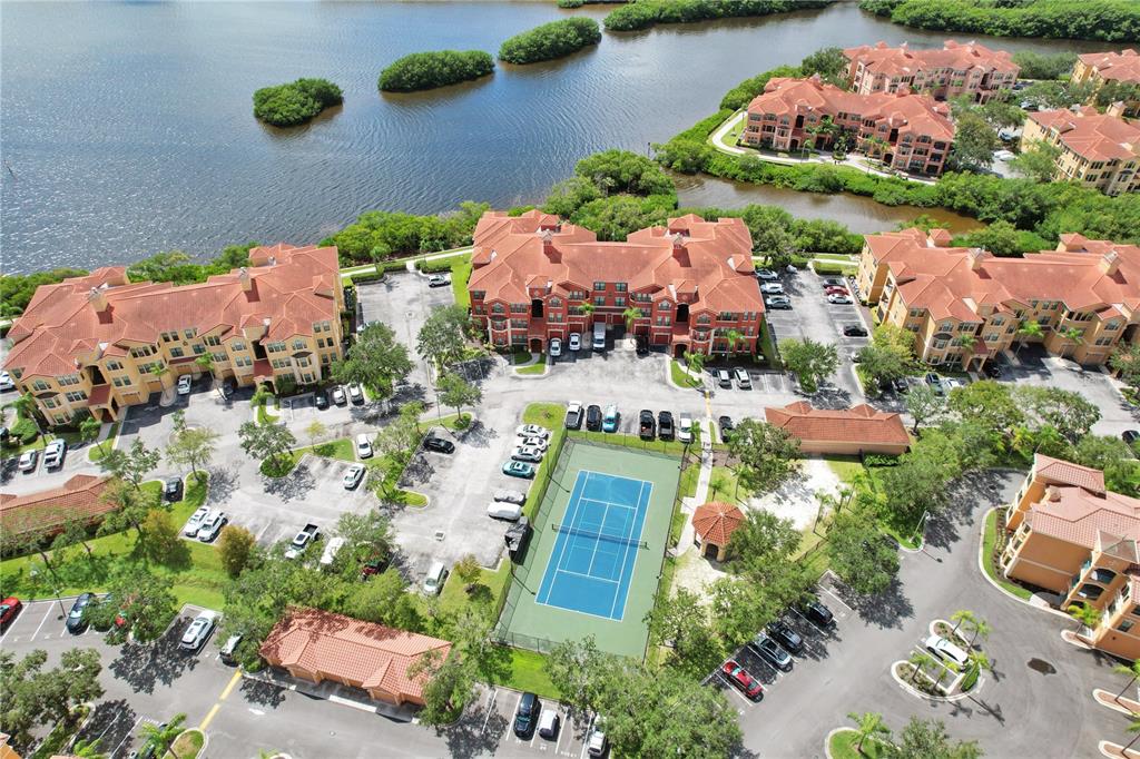 An aerial view shows multi-story apartment buildings, a tennis court, and parking lots near water — 2731 VIA CAPRI UNIT 928, CLEARWATER, FL