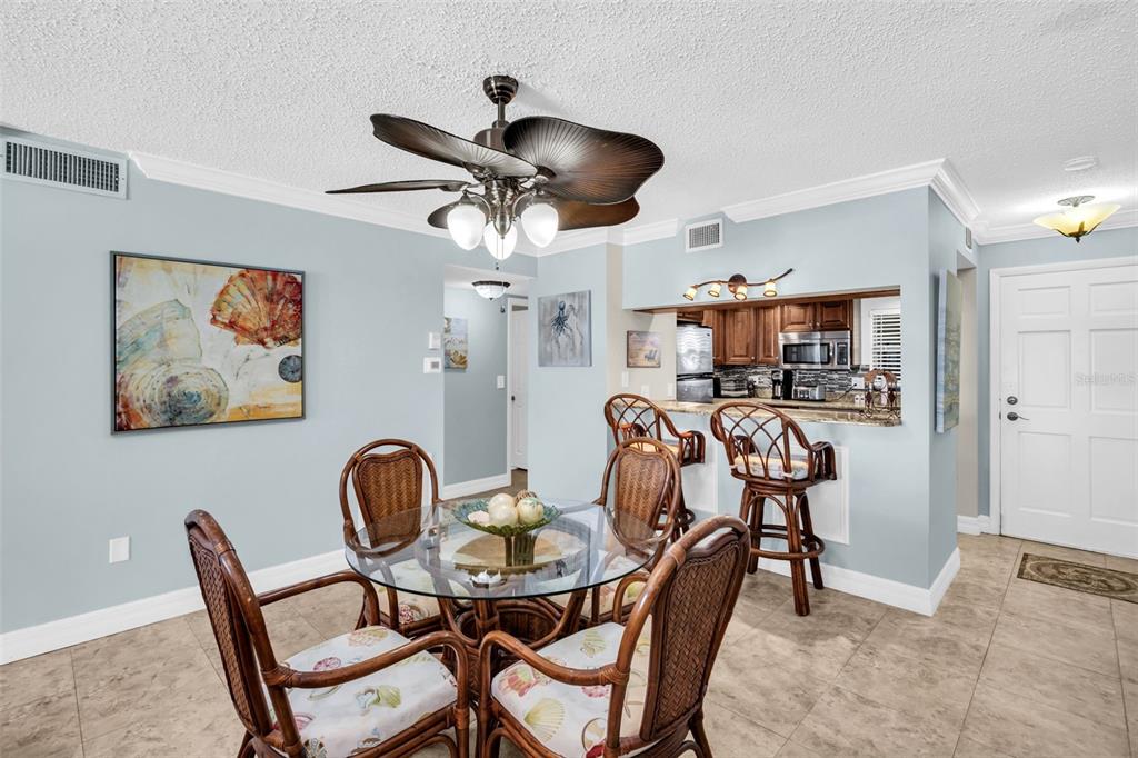 A dining area with a glass table and rattan chairs sits before an open kitchen — 3500 GULF BOULEVARD UNIT 407B, BELLEAIR BEACH, FL