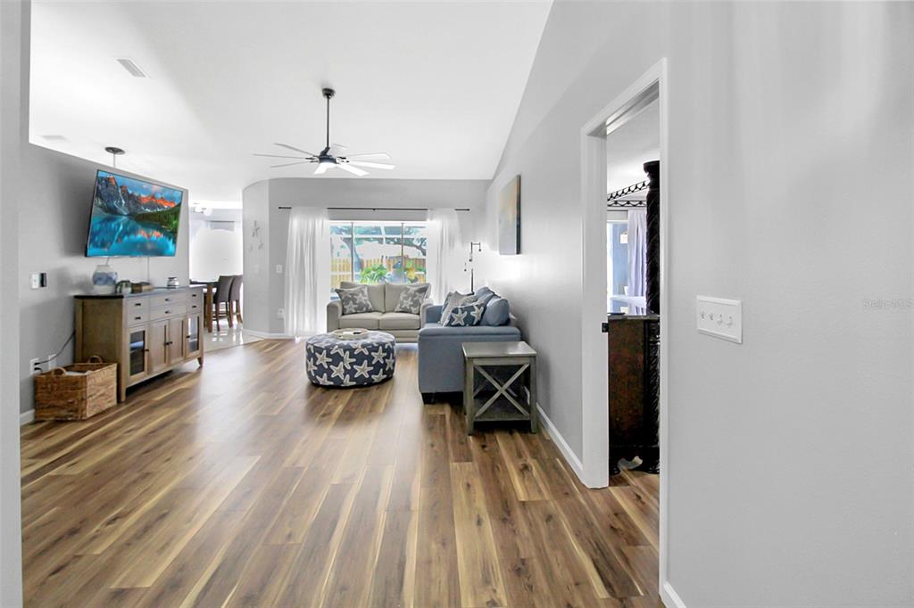 A living room features wood-look flooring, a blue sofa, a patterned ottoman, and a television — 5111 13TH AVENUE DRIVE W, BRADENTON, FL