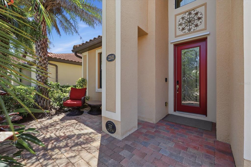 A red front door with a glass insert sits beside a brick patio and chair — 12505 CANAVESE LANE, VENICE, FL