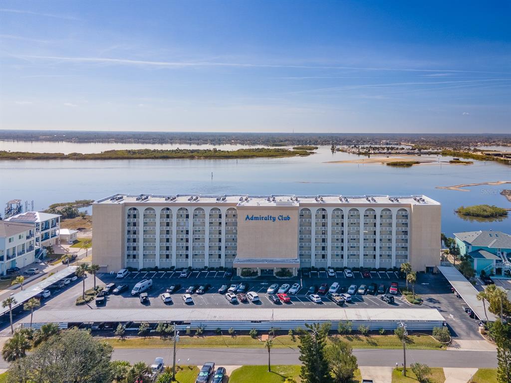 An aerial view shows a multi-story Admiralty Club apartment building with a parking lot and waterfront — 3606 S PENINSULA DRIVE UNIT 809, PORT ORANGE, FL