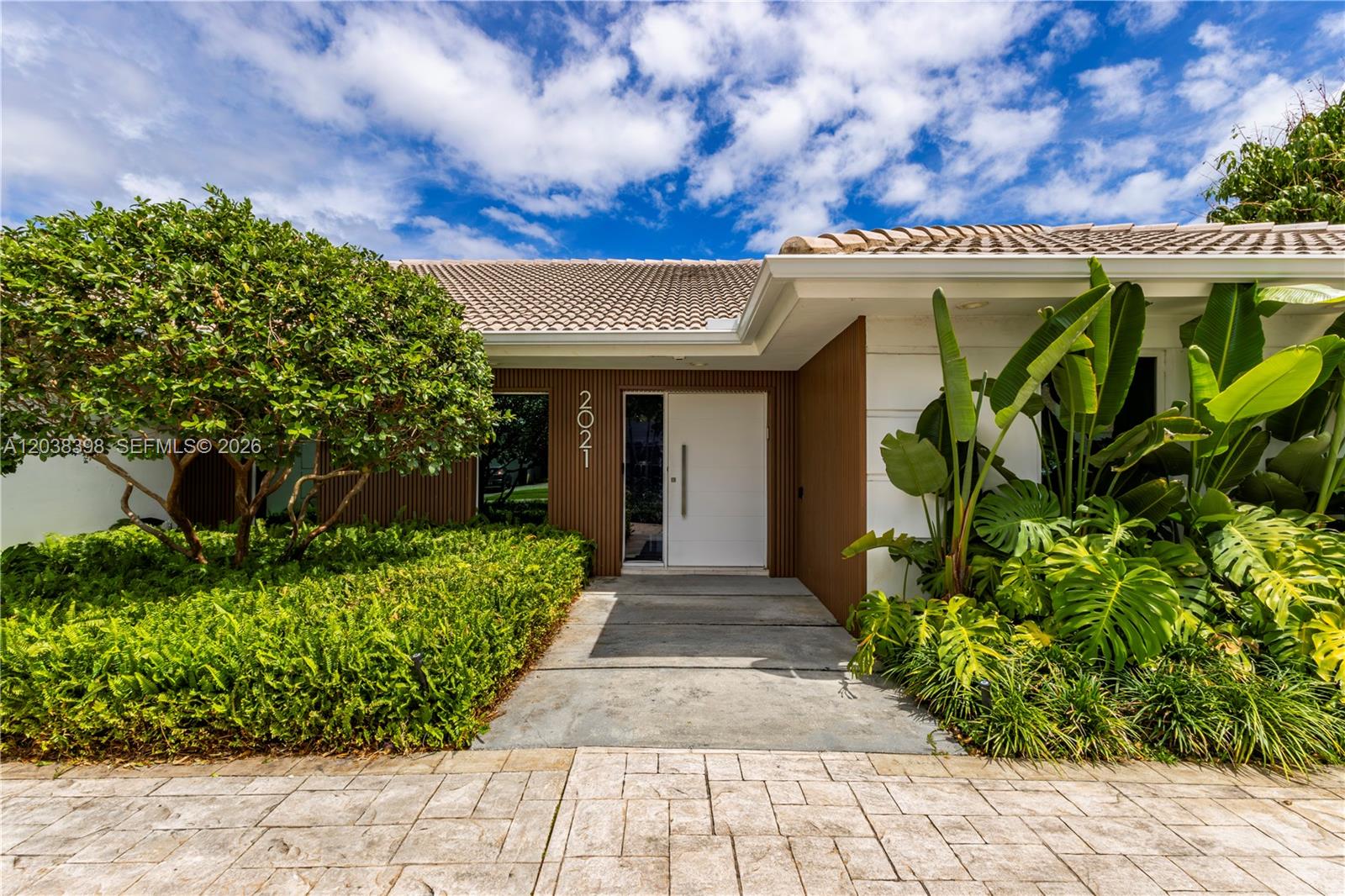 The exterior of a house features a white front door, wood-slat siding, and lush landscaping — 2021 NE 212th St, Miami FL 33179, MIAMI, FL