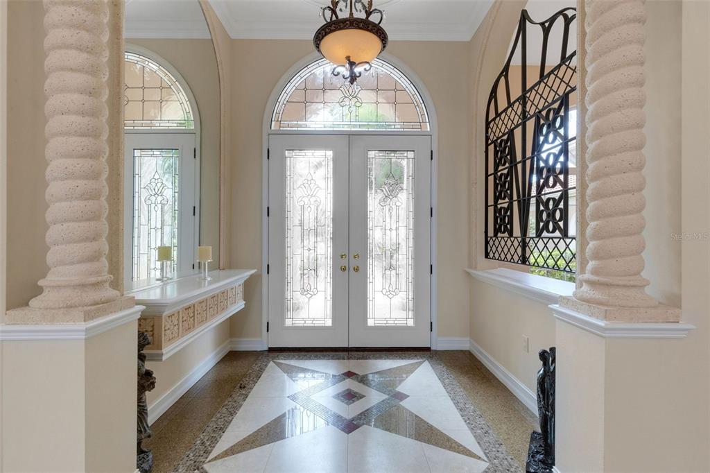 An entryway features double glass doors, stone columns, a patterned tile floor, and a chandelier — 7004 PORTMARNOCK PLACE, BRADENTON, FL