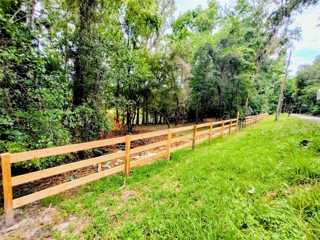 A wooden split-rail fence runs along the edge of a grassy area bordering a forest — FITZHUGH AVENUE, BROOKSVILLE, FL