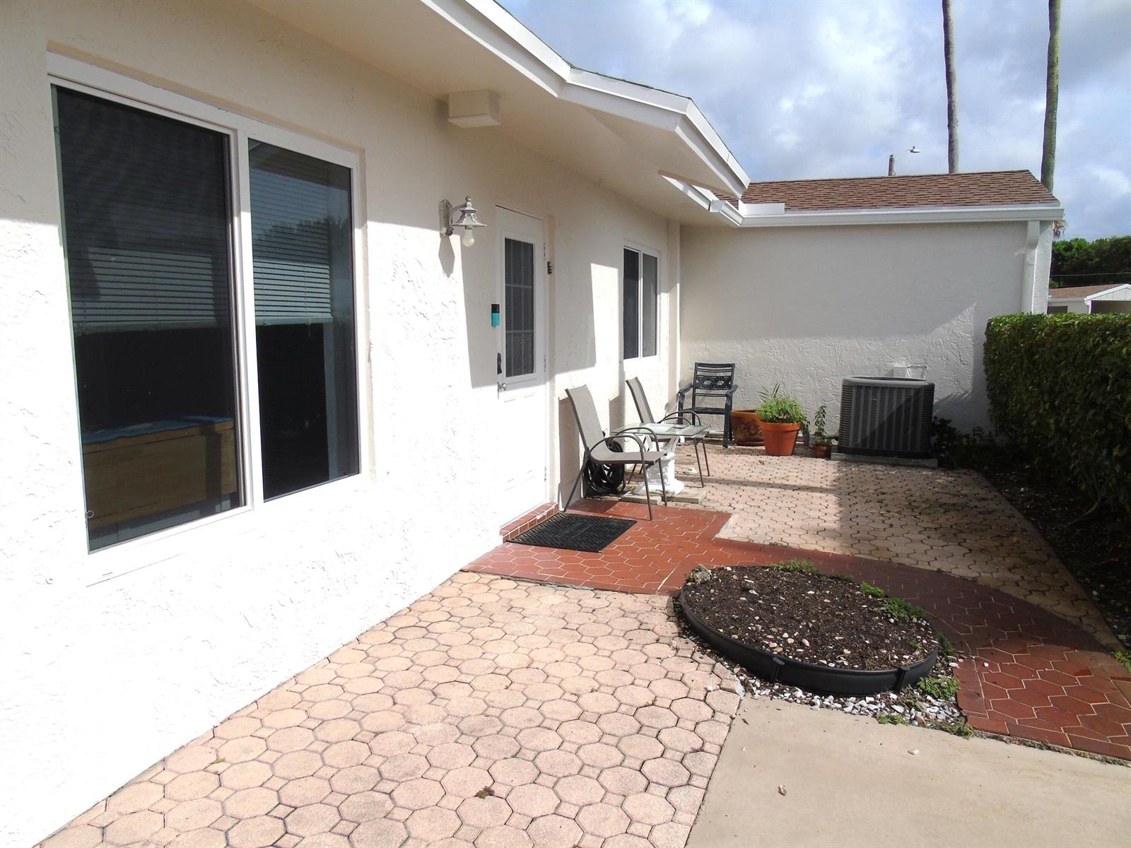 A patio with light-colored pavers and a small garden bed sits outside a white house — 14723 Wildflower Lane, Delray Beach FL 33446, DELRAY BEACH, FL
