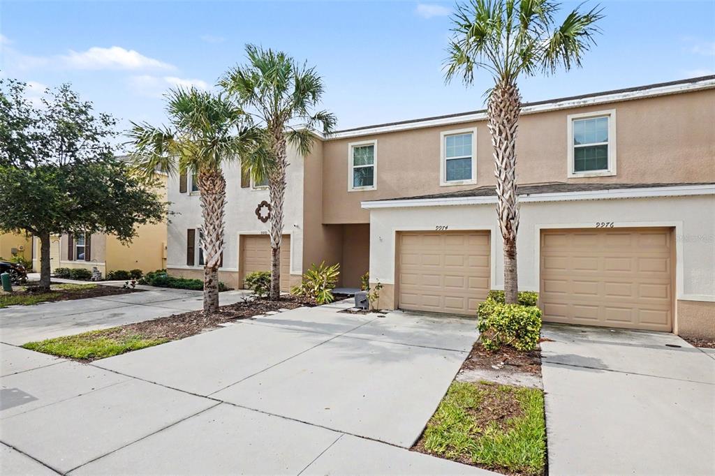 A two-story townhouse exterior features tan stucco walls, two garage doors, and palm trees — 9974 HOUND CHASE DRIVE, GIBSONTON, FL