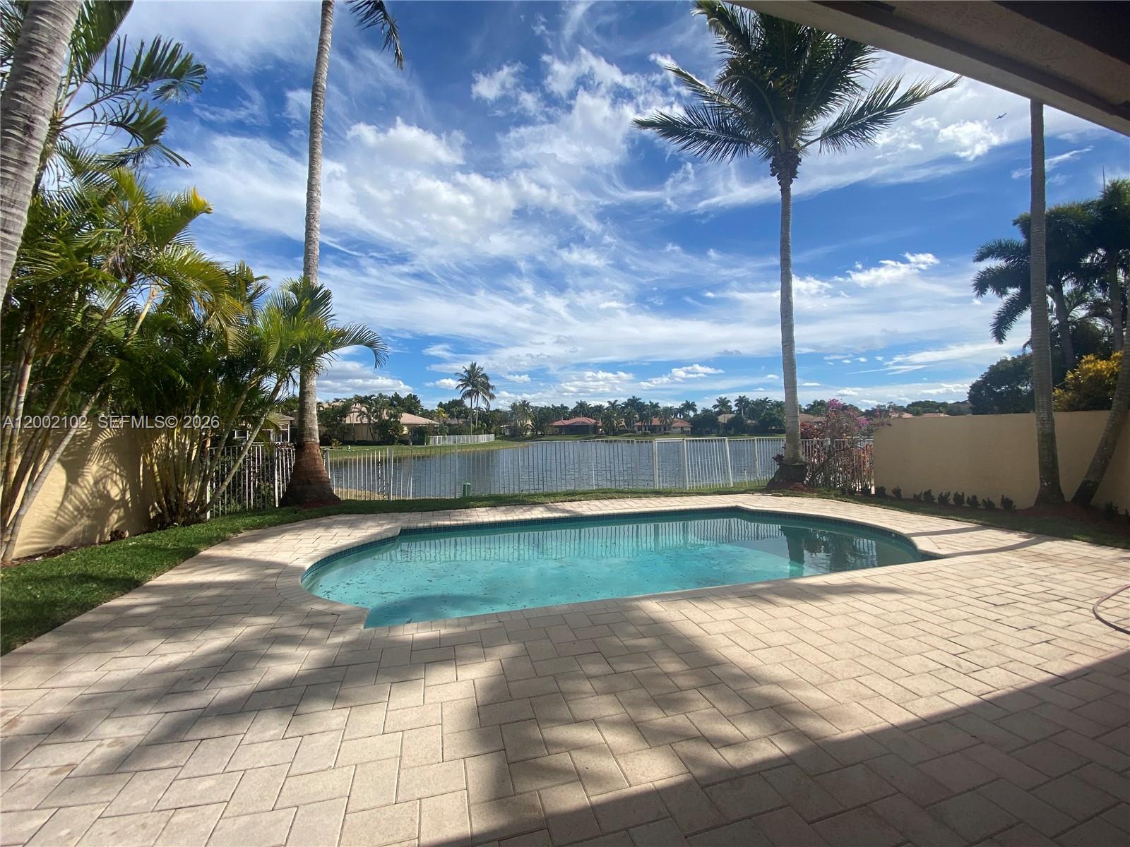 A swimming pool sits on a paved patio overlooking a lake with palm trees — 2486 Bay Isle Dr, Weston FL 33327, WESTON, FL
