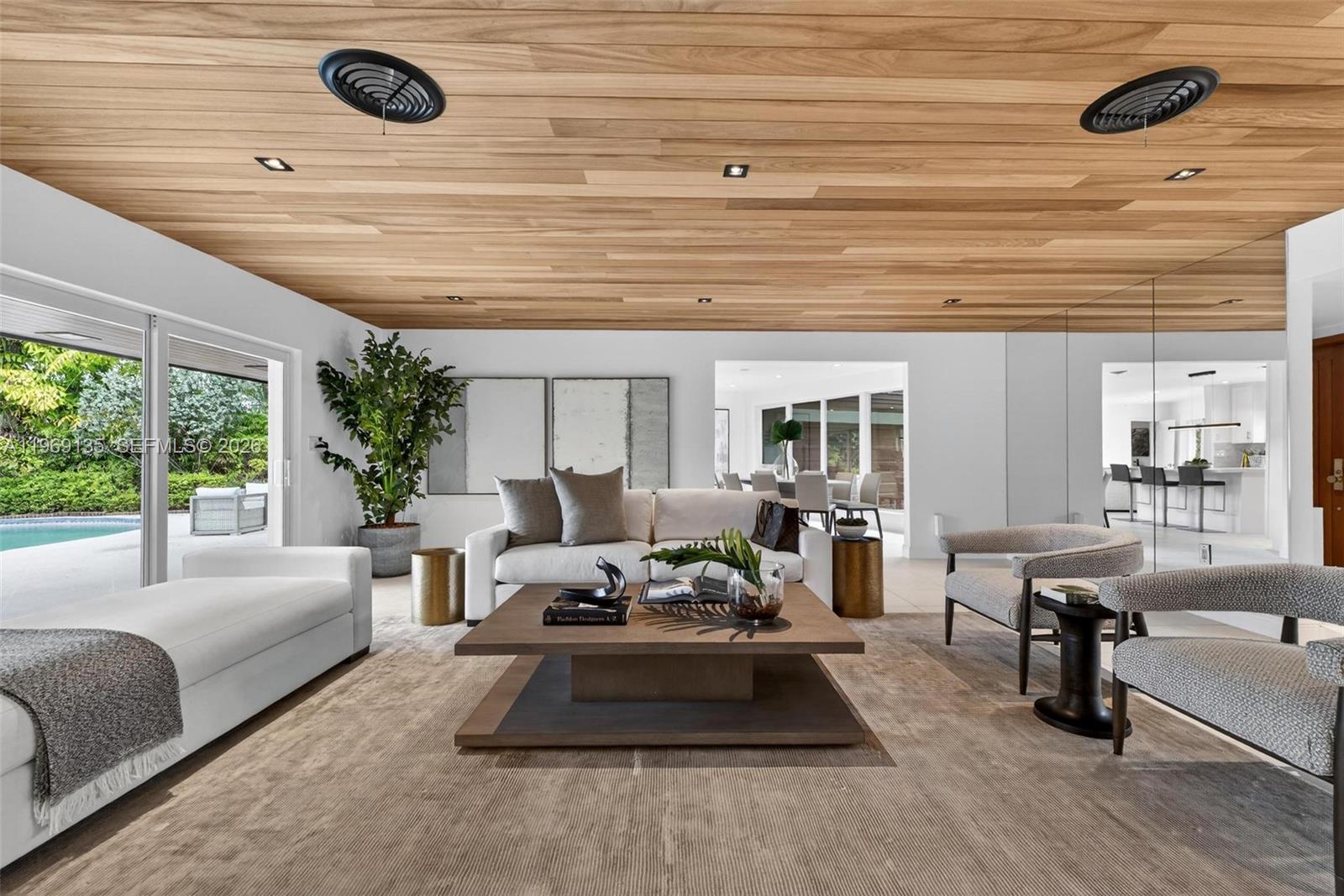 A modern living room features a wood-paneled ceiling, white walls, and a view of a pool — 13060 Lerida St, Coral Gables FL 33156, CORAL GABLES, FL