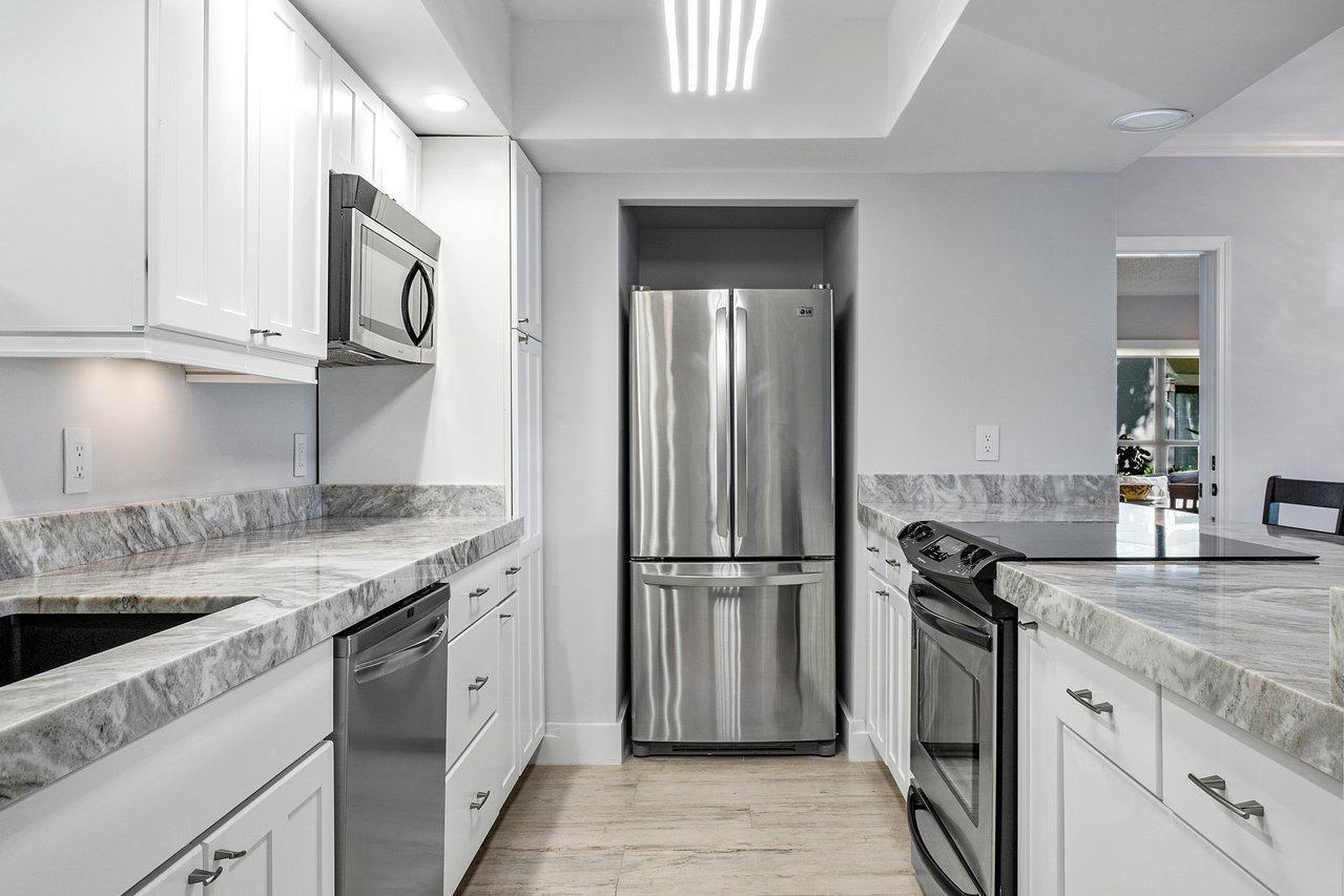 A galley kitchen features white cabinets, stainless steel appliances, and gray-veined stone countertops — 11863 Wimbledon Circle # 445, Wellington FL 33414, WELLINGTON, FL