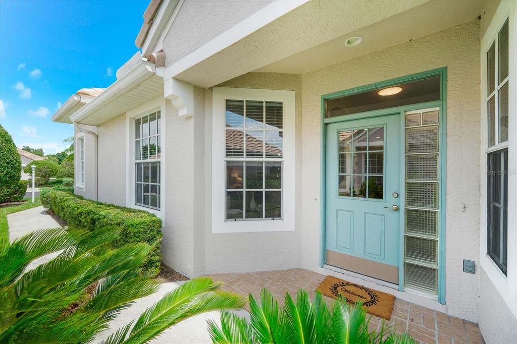 A light blue front door with glass panels sits under a covered entryway with brick flooring — 6942 WOODMORE TERRACE, LAKEWOOD RANCH, FL