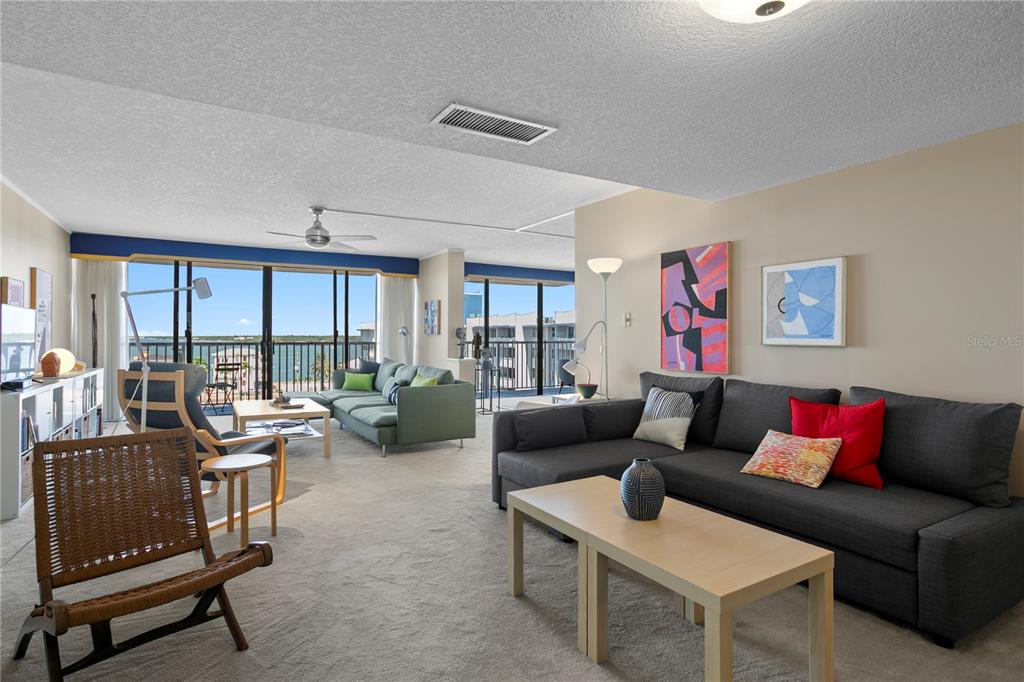 A spacious living room features two gray sectionals, carpeted floors, and a balcony with water views — 450 TREASURE ISLAND CAUSEWAY UNIT 702, TREASURE ISLAND, FL