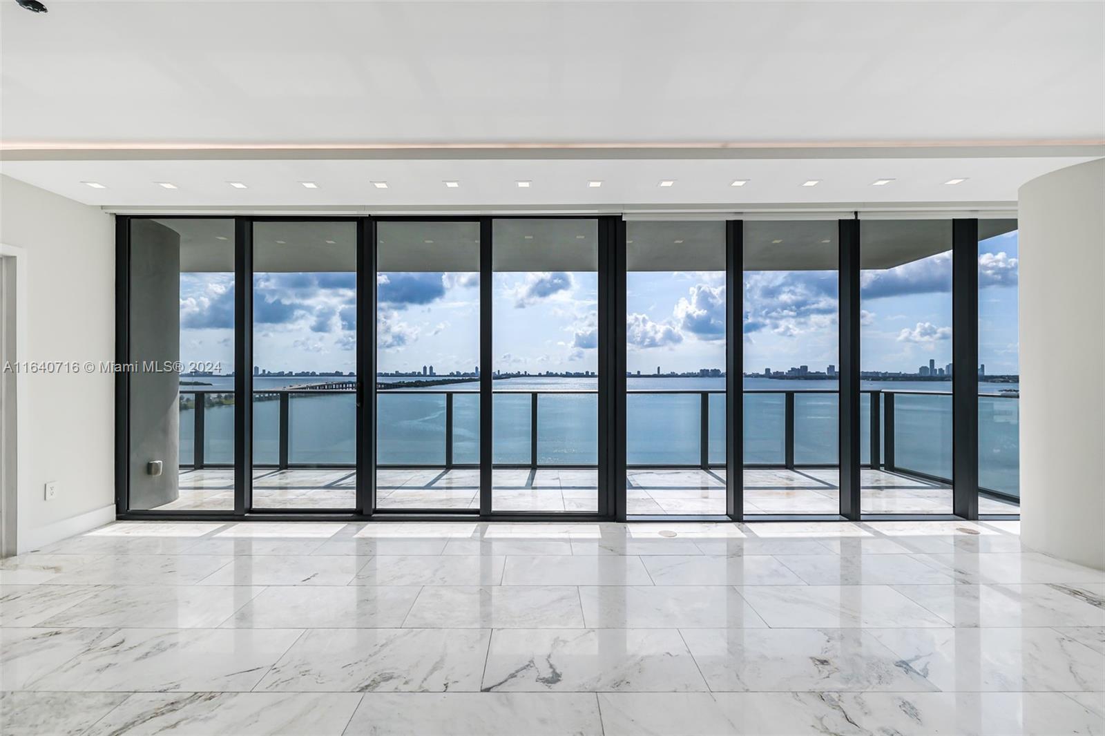 An empty room with white marble floors features floor-to-ceiling windows overlooking a bay and skyline — 3131 NE 7th Ave # 1101, Miami FL 33137, MIAMI, FL