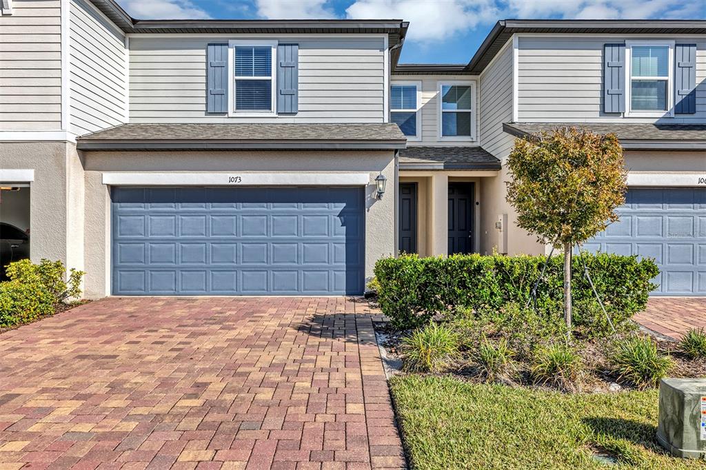 A two-story townhouse exterior features a blue garage door, brick driveway, and manicured landscaping — 1073 DESERT CANDLE DRIVE, OCOEE, FL