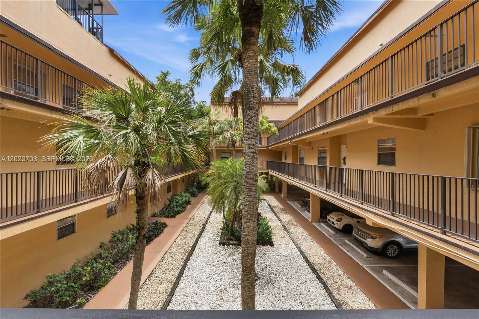 An outdoor courtyard features a gravel garden, palm trees, and covered parking beneath apartment balconies — 430 Commodore Dr # 213, Plantation FL 33325, PLANTATION, FL