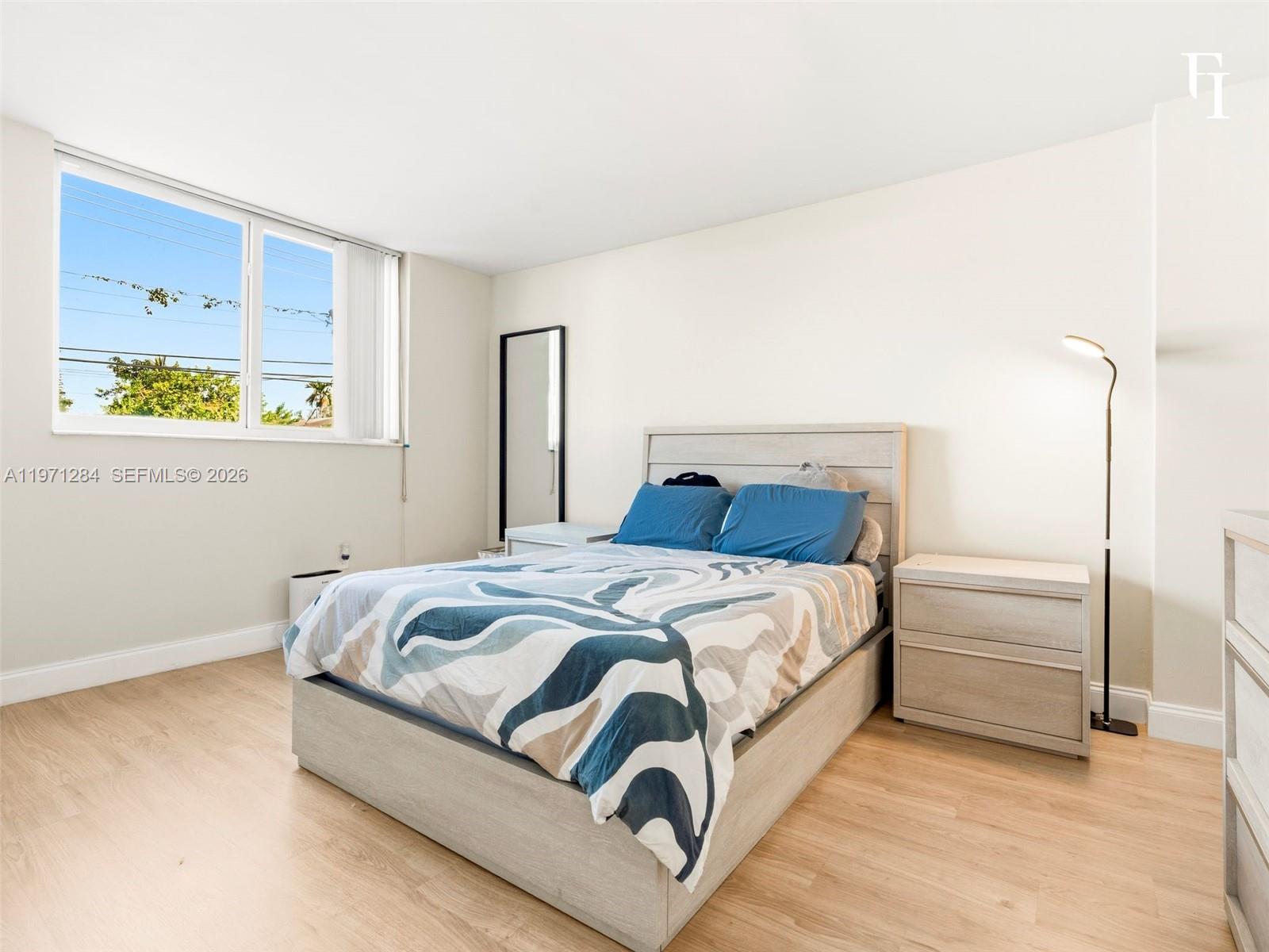 A bedroom features light wood flooring, a bed with a patterned duvet, and a floor lamp — 1251 NE 108th St # 108, Miami FL 33161, MIAMI, FL